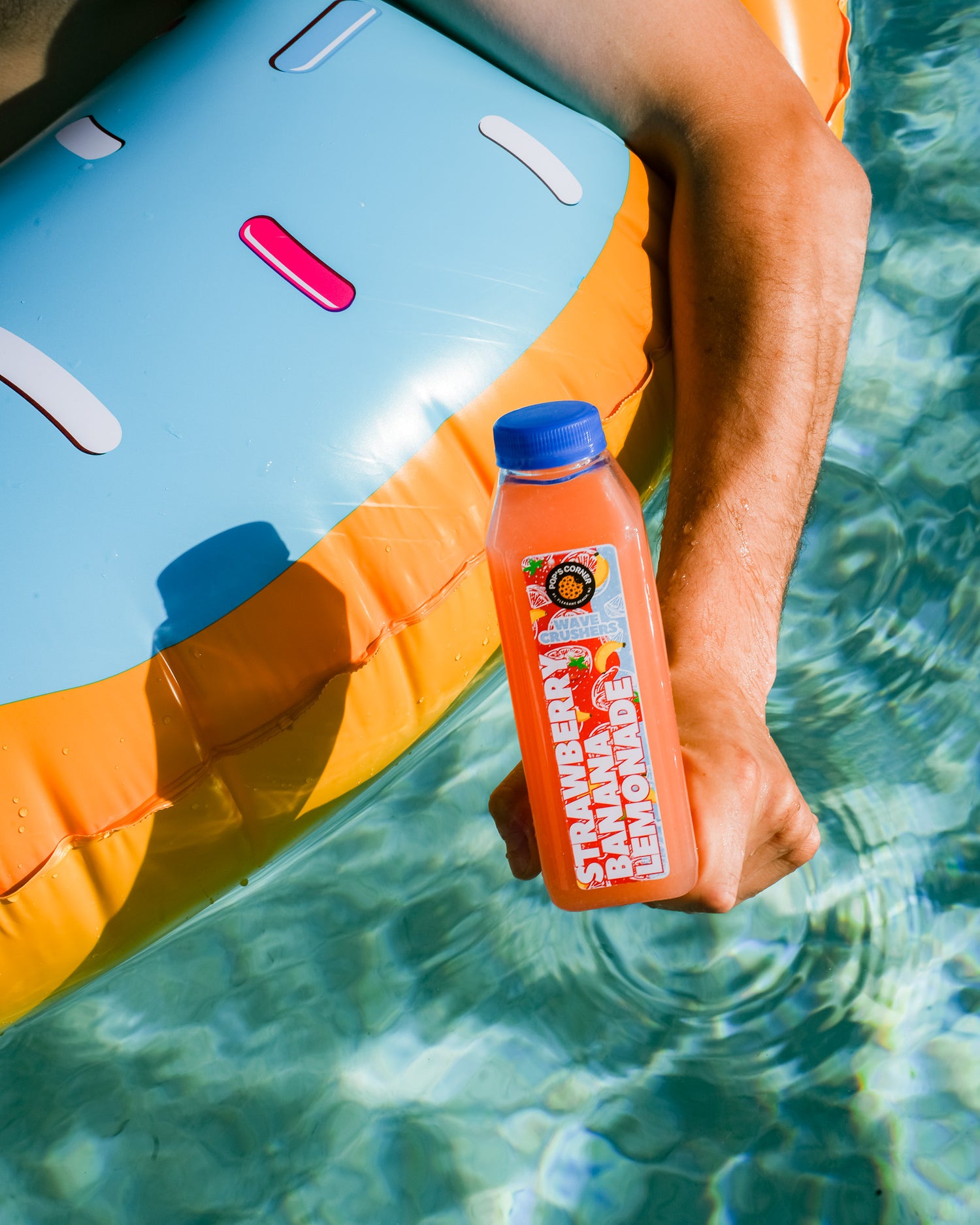 Person holding a strawberry banana lemonade bottle on an inflatable pool float in clear water.